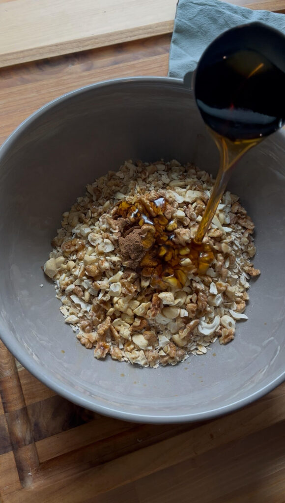 Rolled oats in large bowl with chopped nuts, maple syrup, and coconut oil.