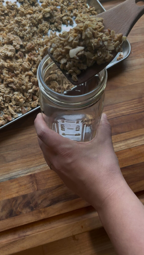 Homemade granola being put into large mouth mason jar by a wooden spatula 